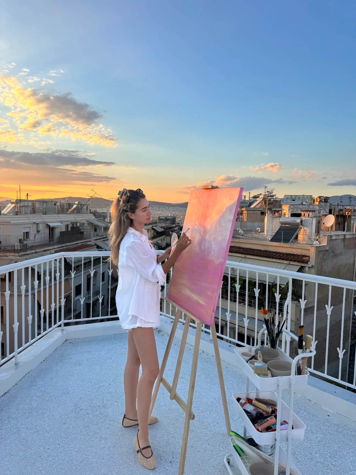Jeanne Richterich painting a soft pink sky canvas on an easel, overlooking Athens rooftops at sunset.