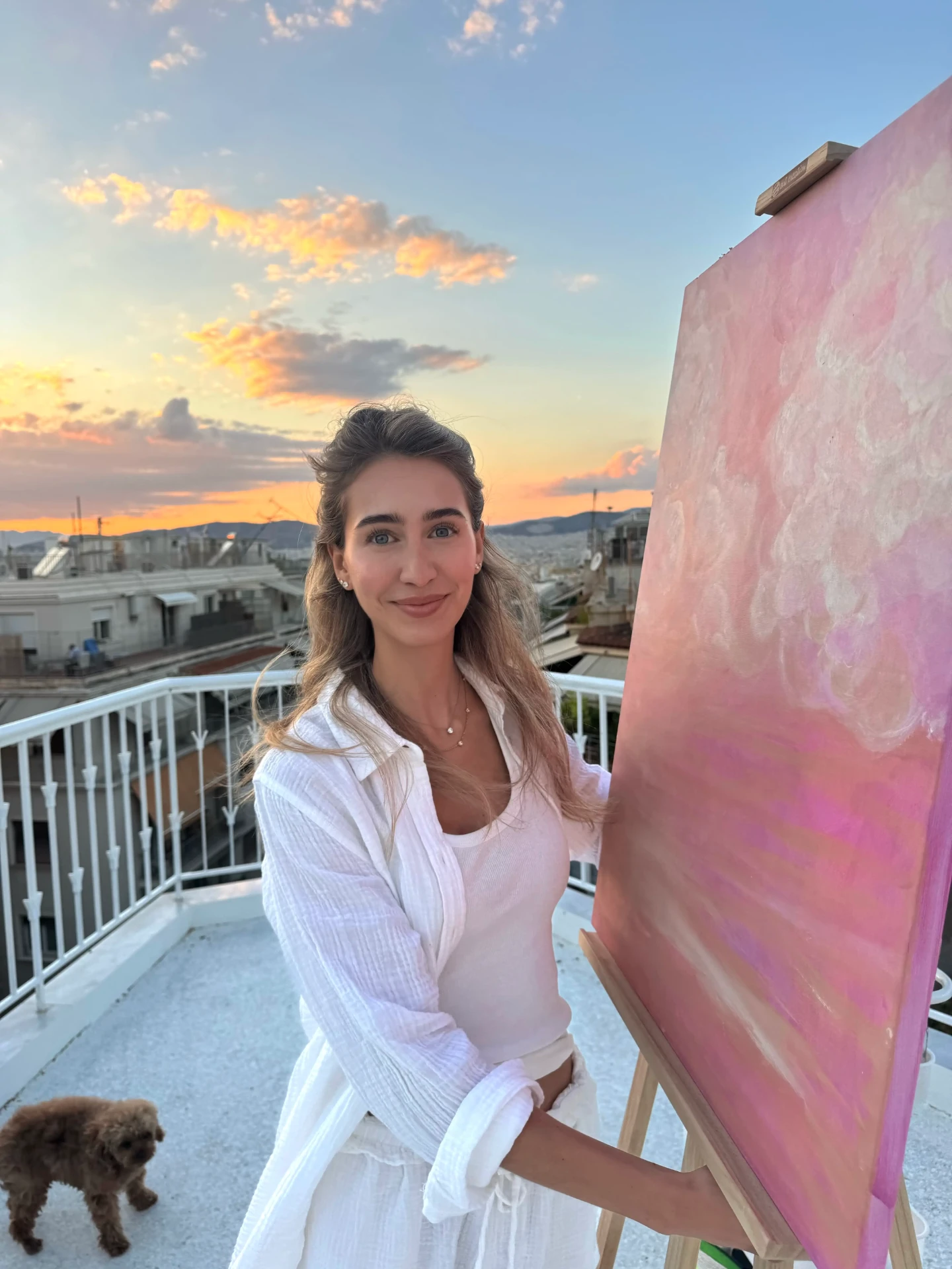 Jeanne Richterich smiling beside a pink canvas on an easel, with her small brown dog on a rooftop at sunset.